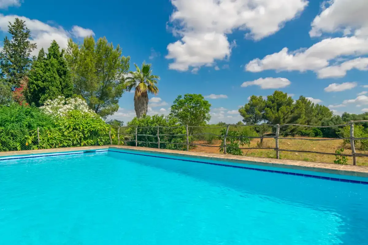 A turquoise swimming pool sits beside a fenced garden under a blue sky.