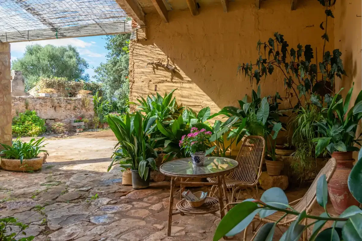A shaded stone patio with potted plants, wicker seating, and a small table.