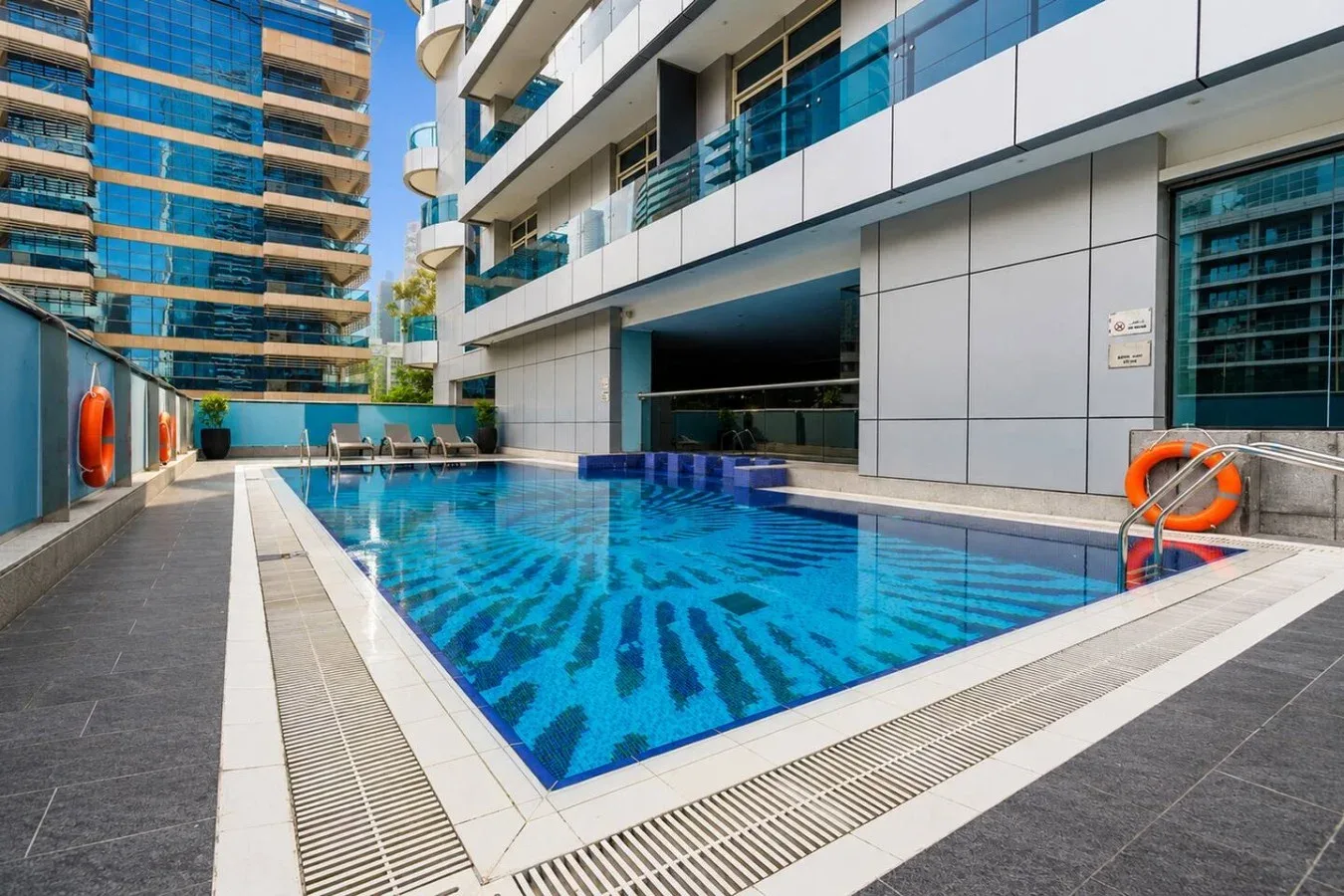 Una piscina al aire libre junto a edificios modernos, con asientos de descanso en el borde.