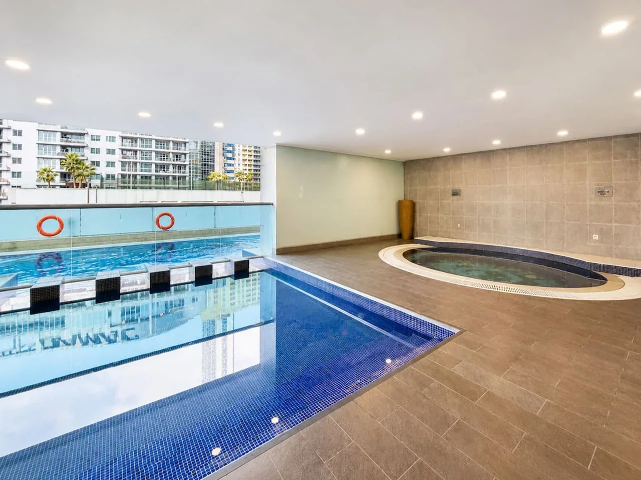 Una piscina cubierta junto a una piscina circular de hidromasaje, con ventanales que dan a edificios cercanos.