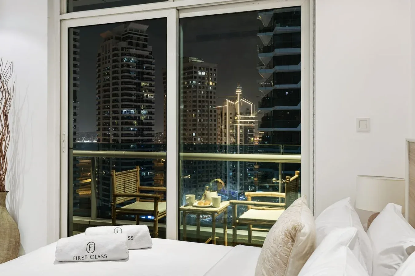 Un dormitorio de noche tiene una gran ventana con vistas a edificios iluminados de la ciudad.