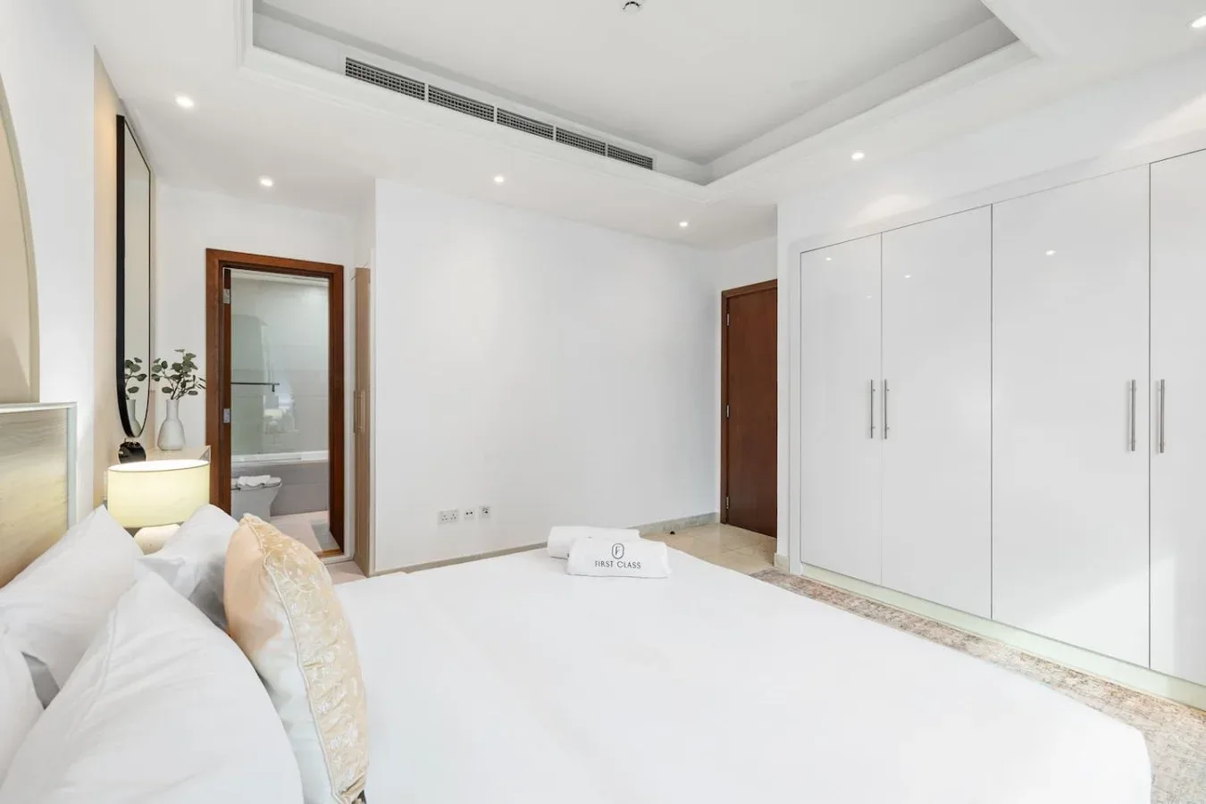 Un dormitorio luminoso con una cama blanca, armarios empotrados y una puerta hacia el baño.