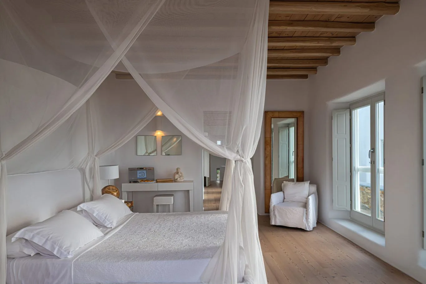 A bright bedroom with a canopy bed and a seating area beside the windows.