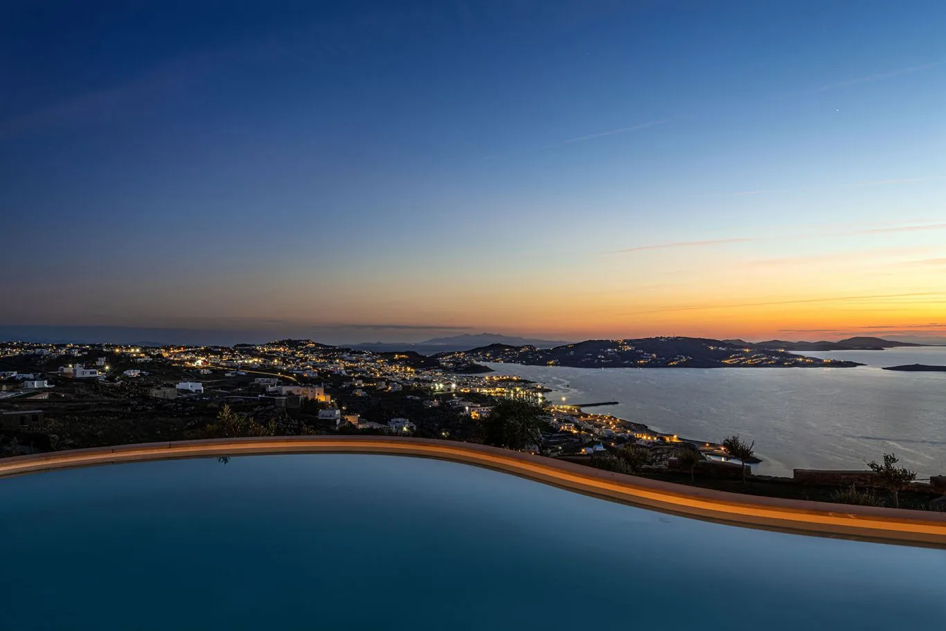 An infinity pool overlooks a coastal town and bay at sunset.
