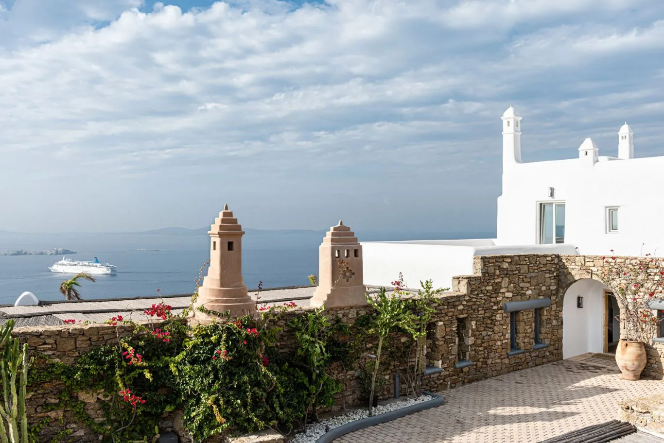 A white seaside building stands beside a stone wall and flower garden overlooking the water.