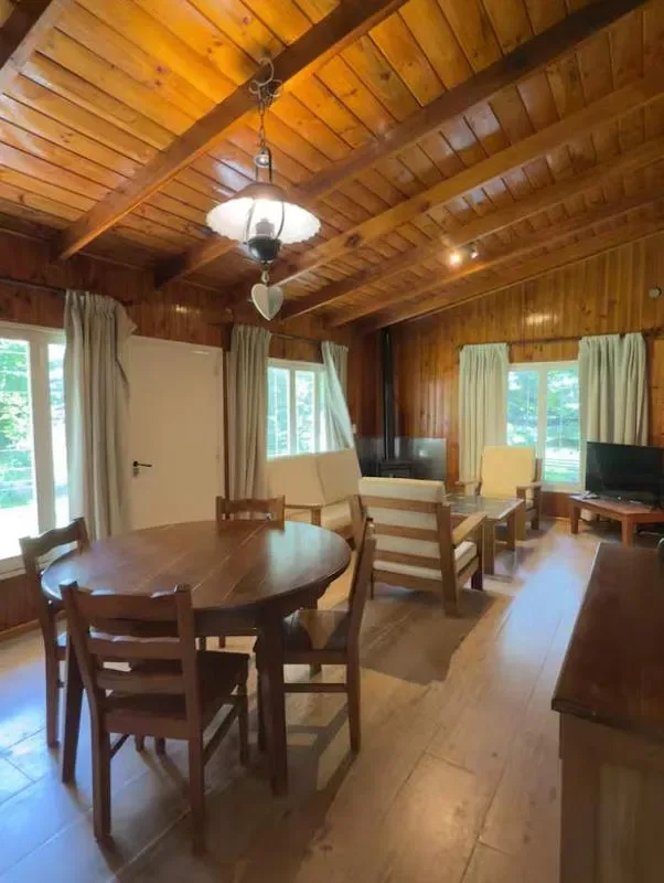 Un comedor y sala revestidos de madera, con una mesa redonda y ventanas grandes.