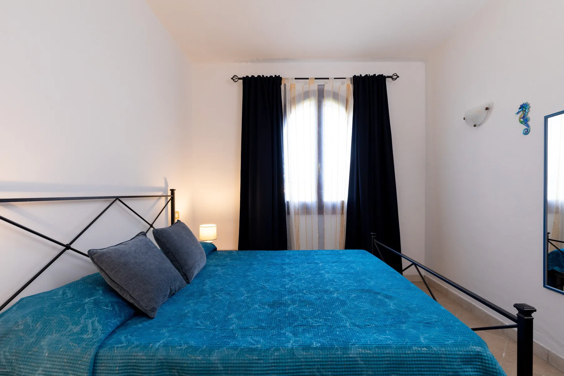 Un dormitorio con una cama cubierta de ropa de cama azul bajo una ventana con cortinas.