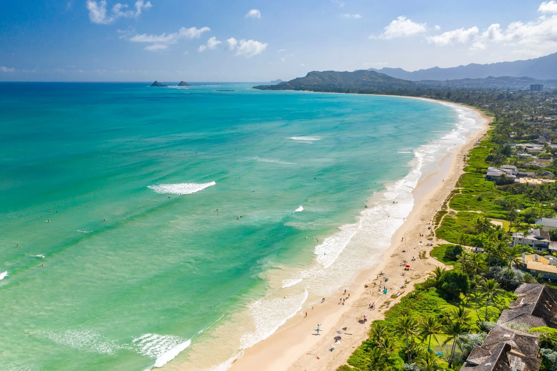Aerial view of a sandy beach curving along turquoise water and coastal greenery.