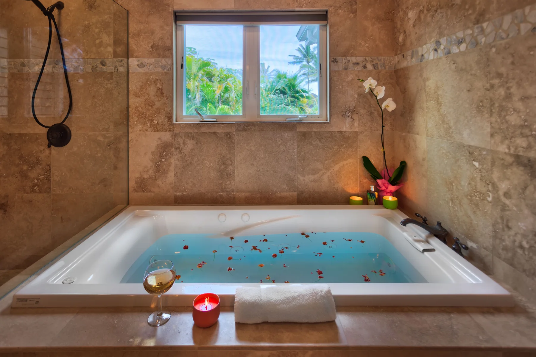 A tiled bathroom features a soaking tub filled with water and flower petals.