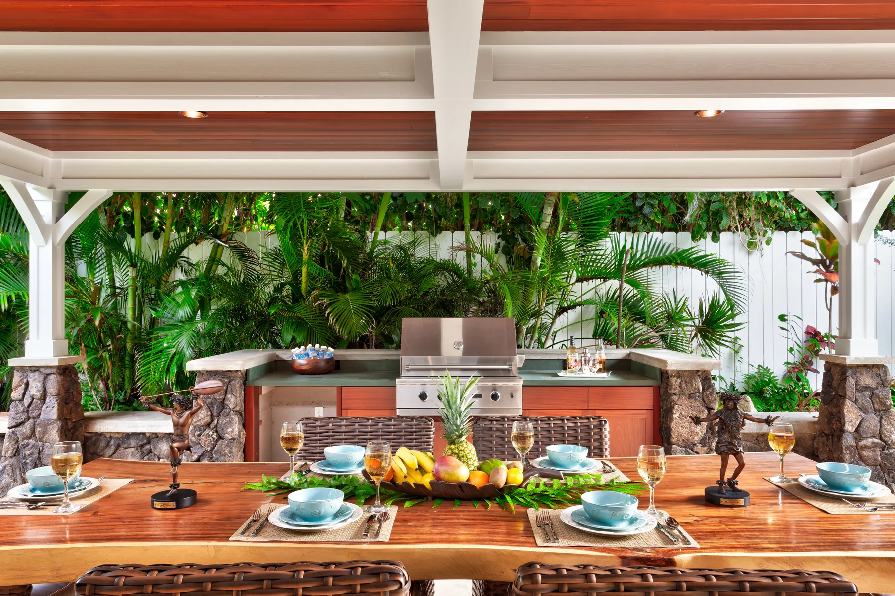 A covered outdoor dining area has a long wooden table set with dishes and tropical fruit.