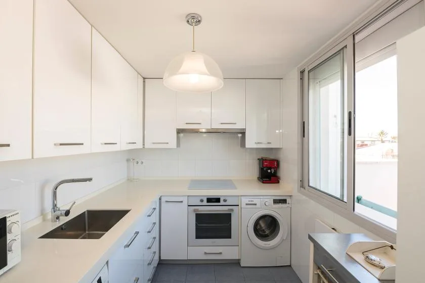 Una cocina blanca y luminosa con horno, lavadora y grandes ventanas.