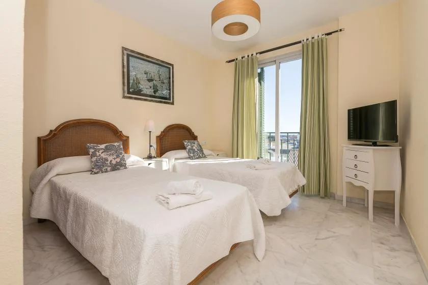 Un dormitorio luminoso con dos camas, cortinas claras y un televisor sobre una cómoda.
