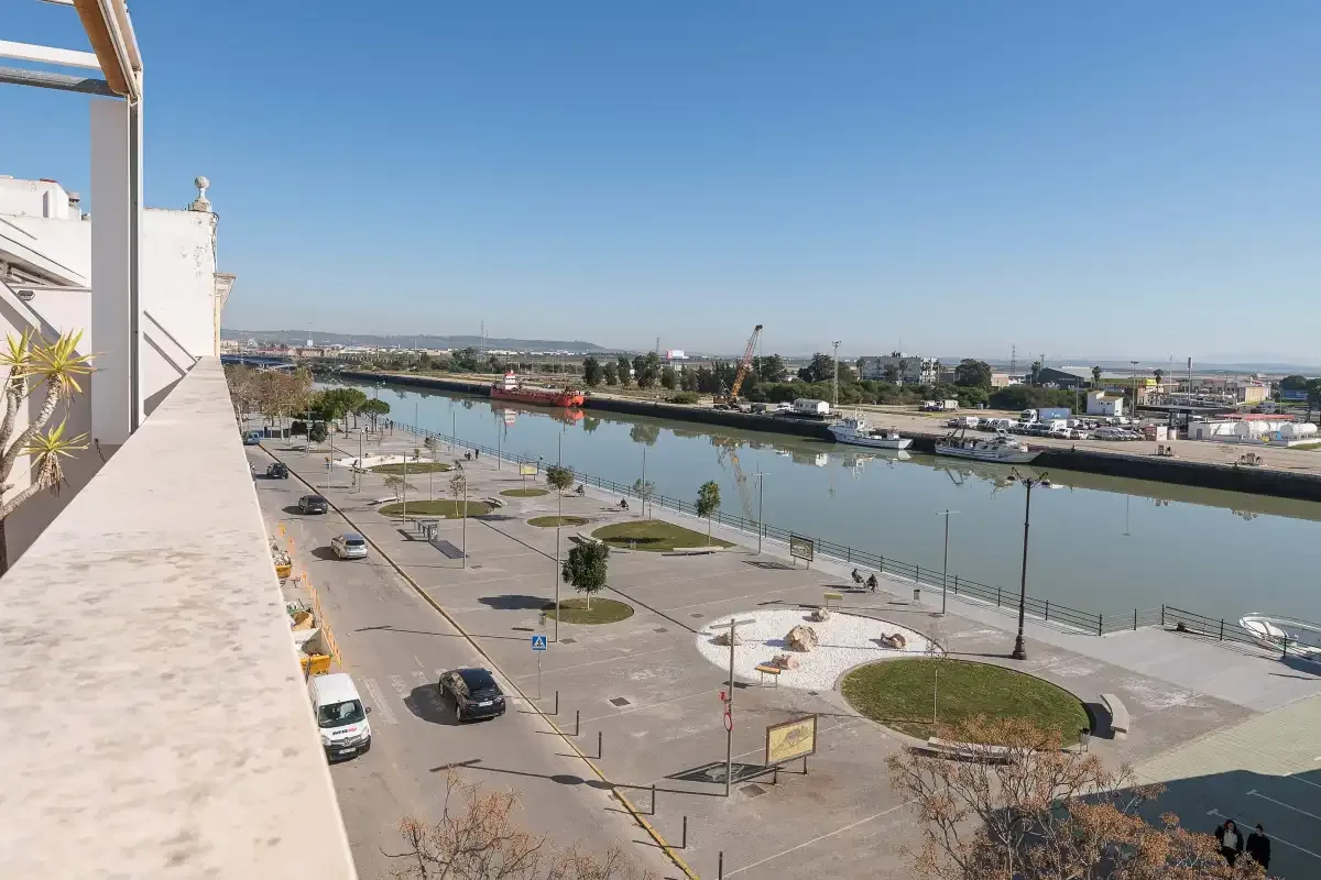 Un balcón da a un paseo junto al agua, una zona de estacionamiento y un río.