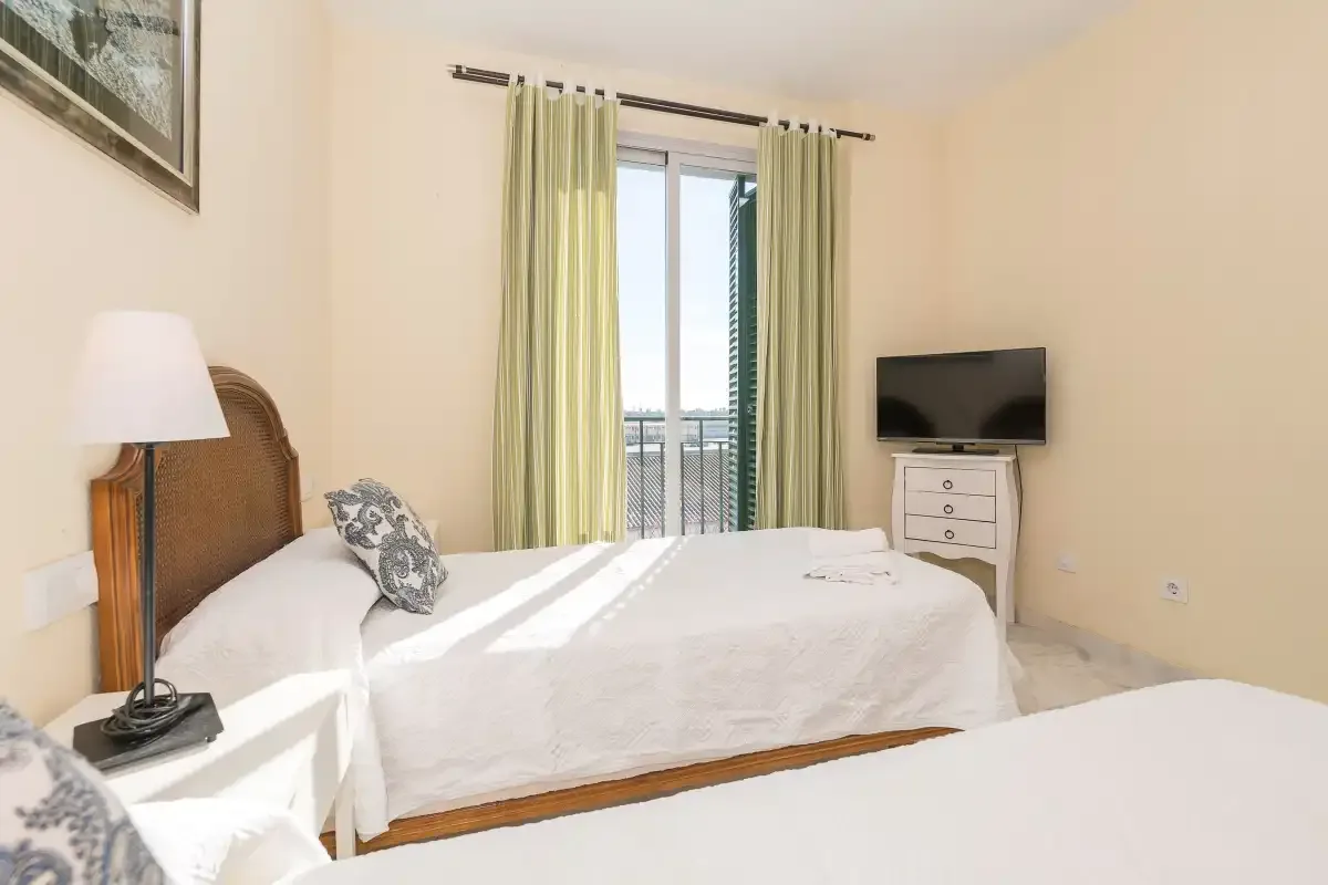 Un dormitorio con una cama, cortinas claras y un televisor.