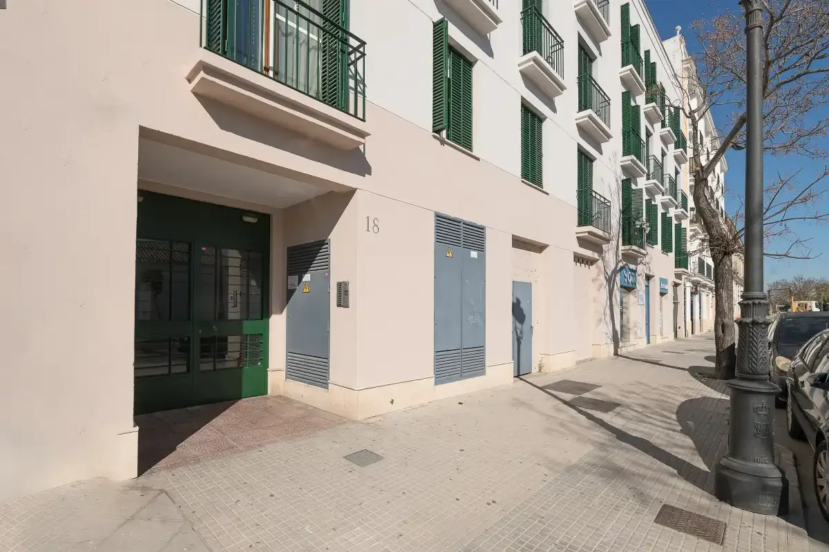 Una acera urbana bordea un edificio claro con balcones verdes.