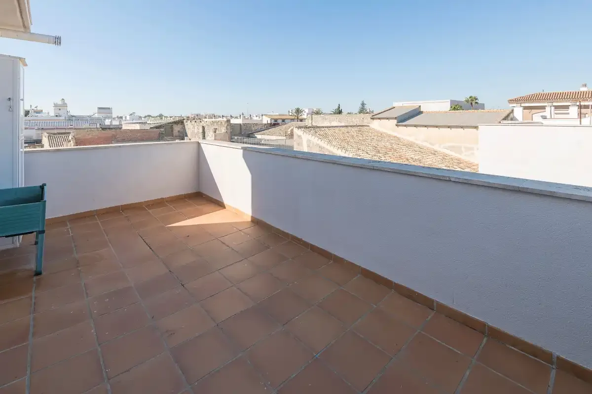 Una terraza soleada con suelo de baldosas y un muro blanco bajo da a los tejados cercanos.