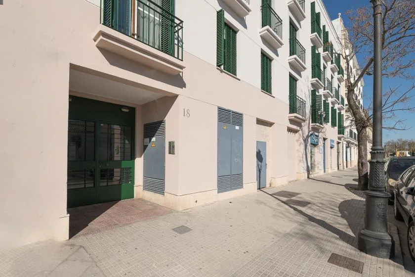 Una acera urbana soleada bordea un edificio blanco con persianas verdes.
