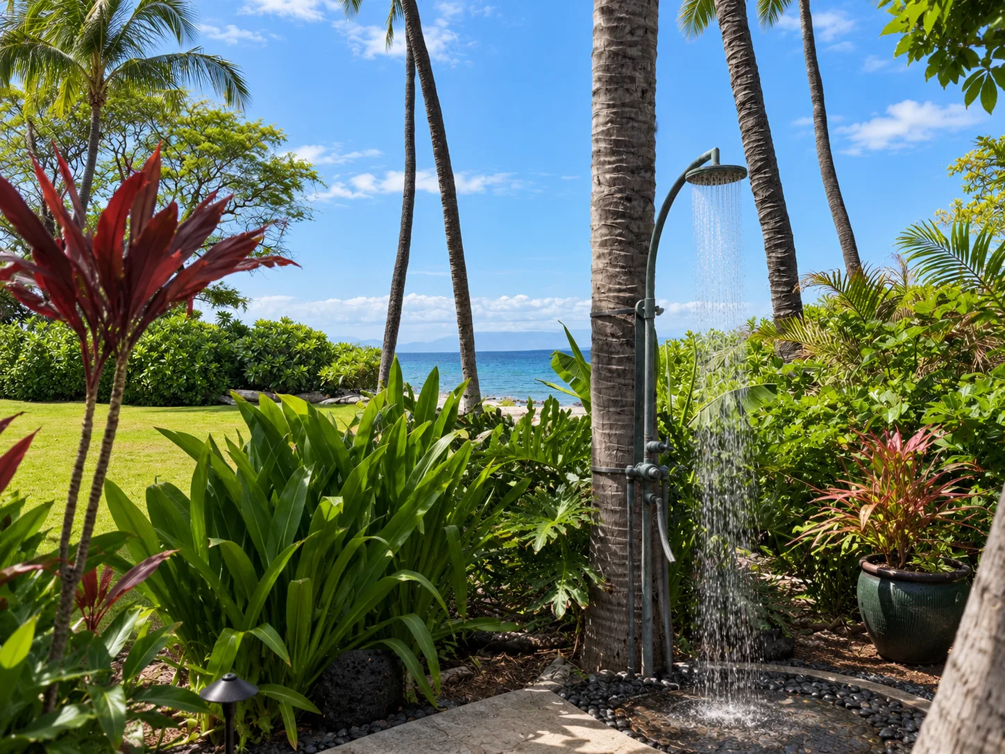 Una ducha exterior está entre plantas tropicales, junto a un jardín con césped y el océano.