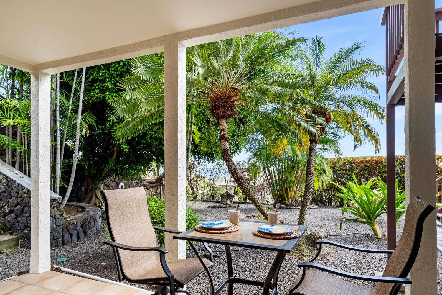 Un patio cubierto tiene una mesa de comedor y asientos con vista a un jardín con palmeras.