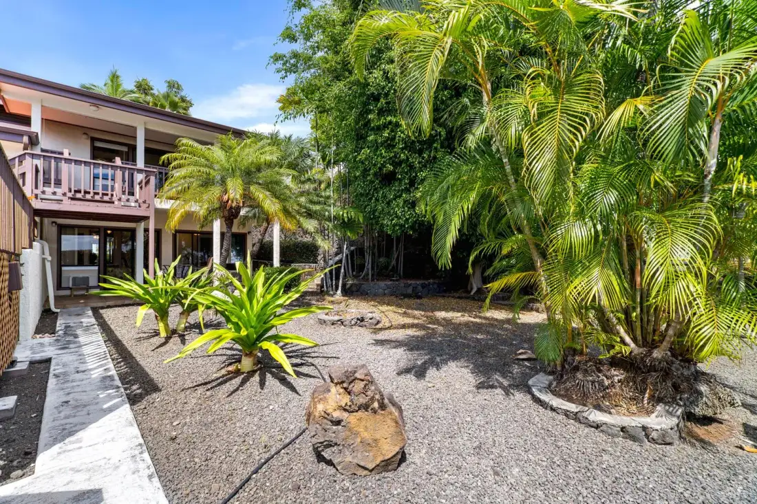 Un patio de grava junto a un edificio de dos pisos está rodeado de palmeras y vegetación tropical.
