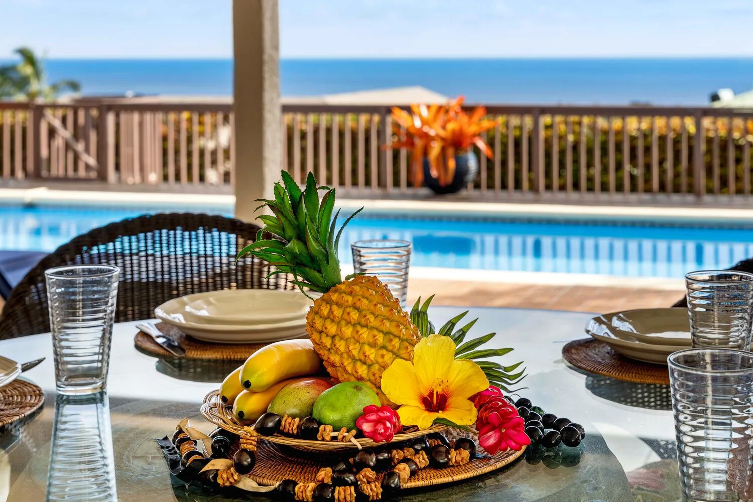 Una mesa de comedor con una bandeja de frutas está junto a una piscina con vista al océano.