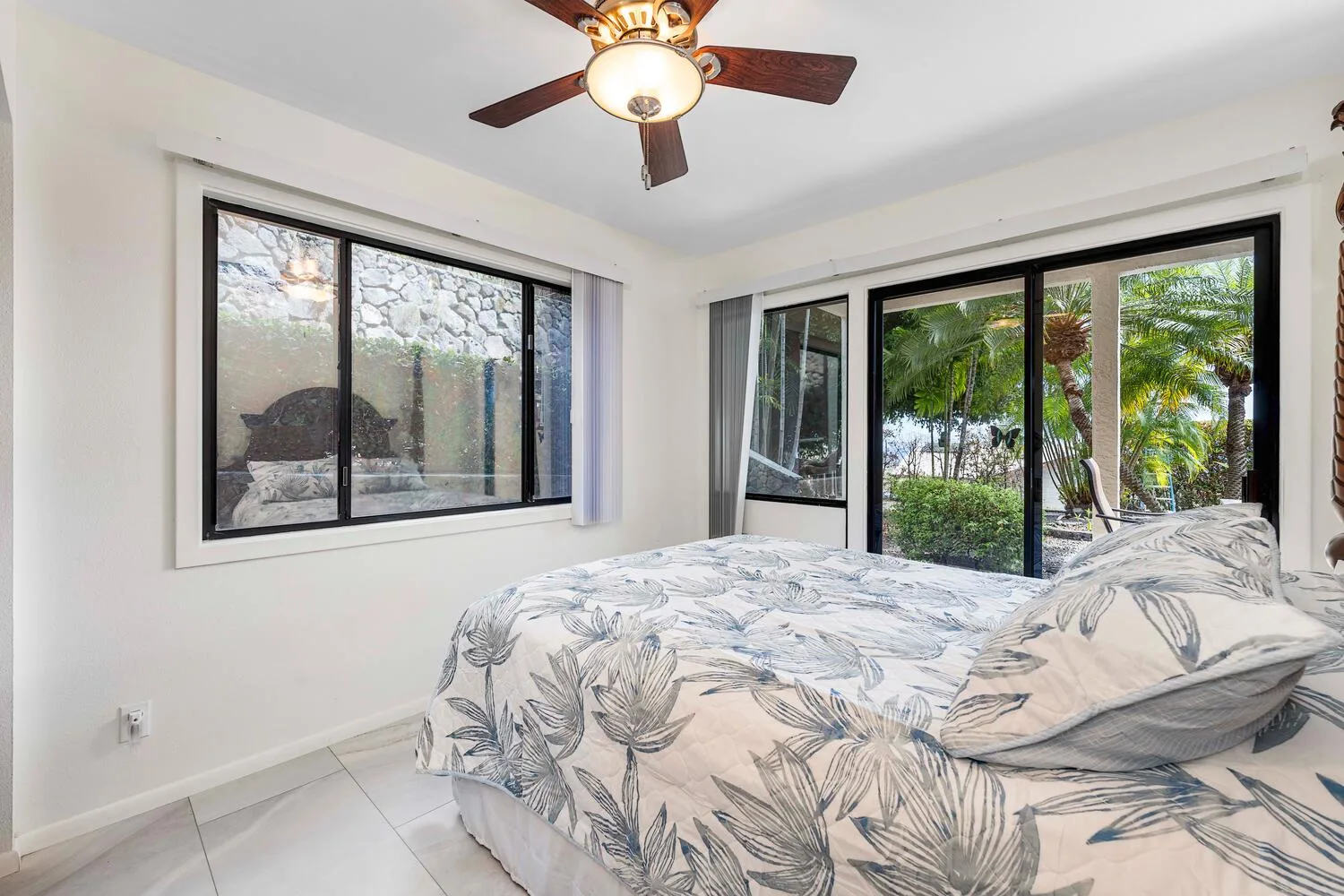 Un dormitorio luminoso con una cama junto a grandes ventanas con vistas a palmeras.