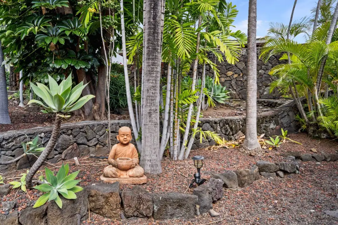 Un jardín tropical presenta una estatua sentada entre palmeras y muros de piedra.