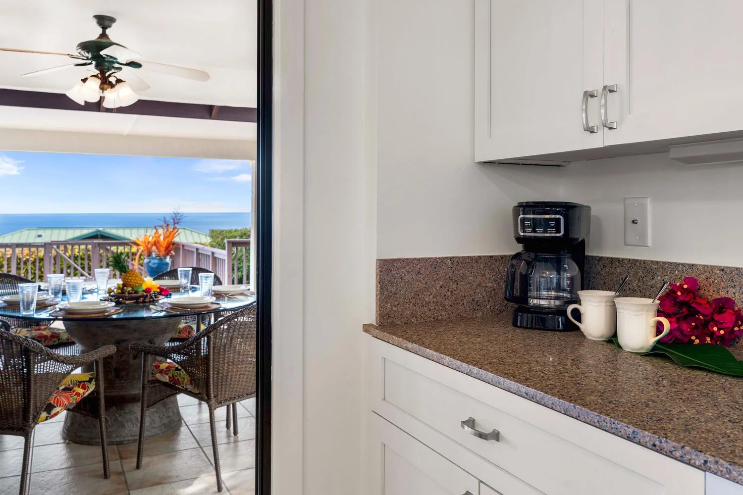 Una encimera de cocina con cafetera y tazas da a un comedor en el balcón y al mar.