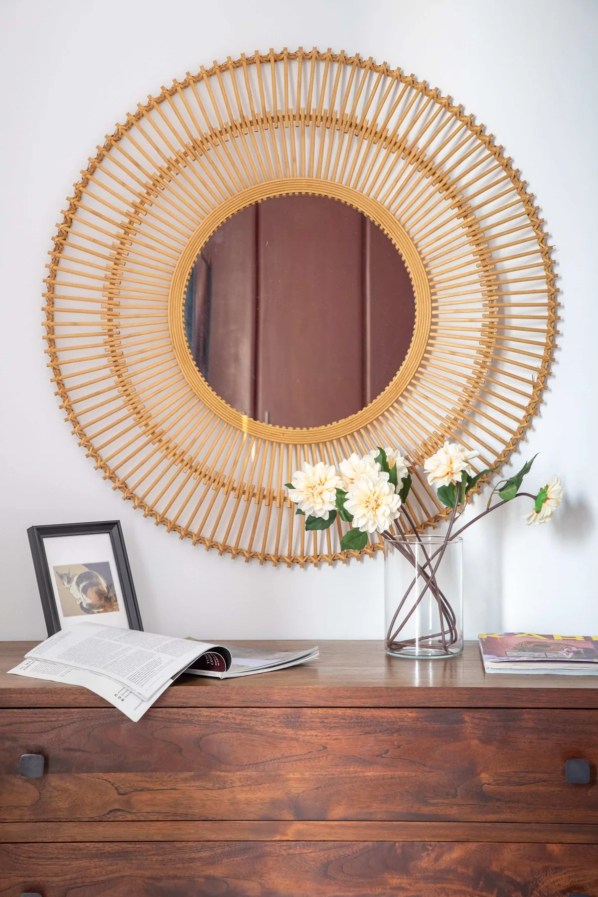 Una cómoda de madera muestra un espejo redondo tejido, flores y un libro abierto.