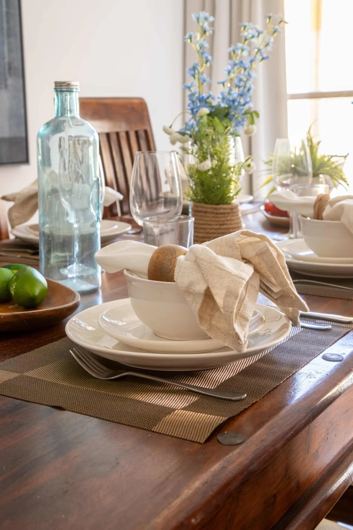 Una mesa de comedor está puesta con platos, tazas, servilletas y un jarrón con flores.