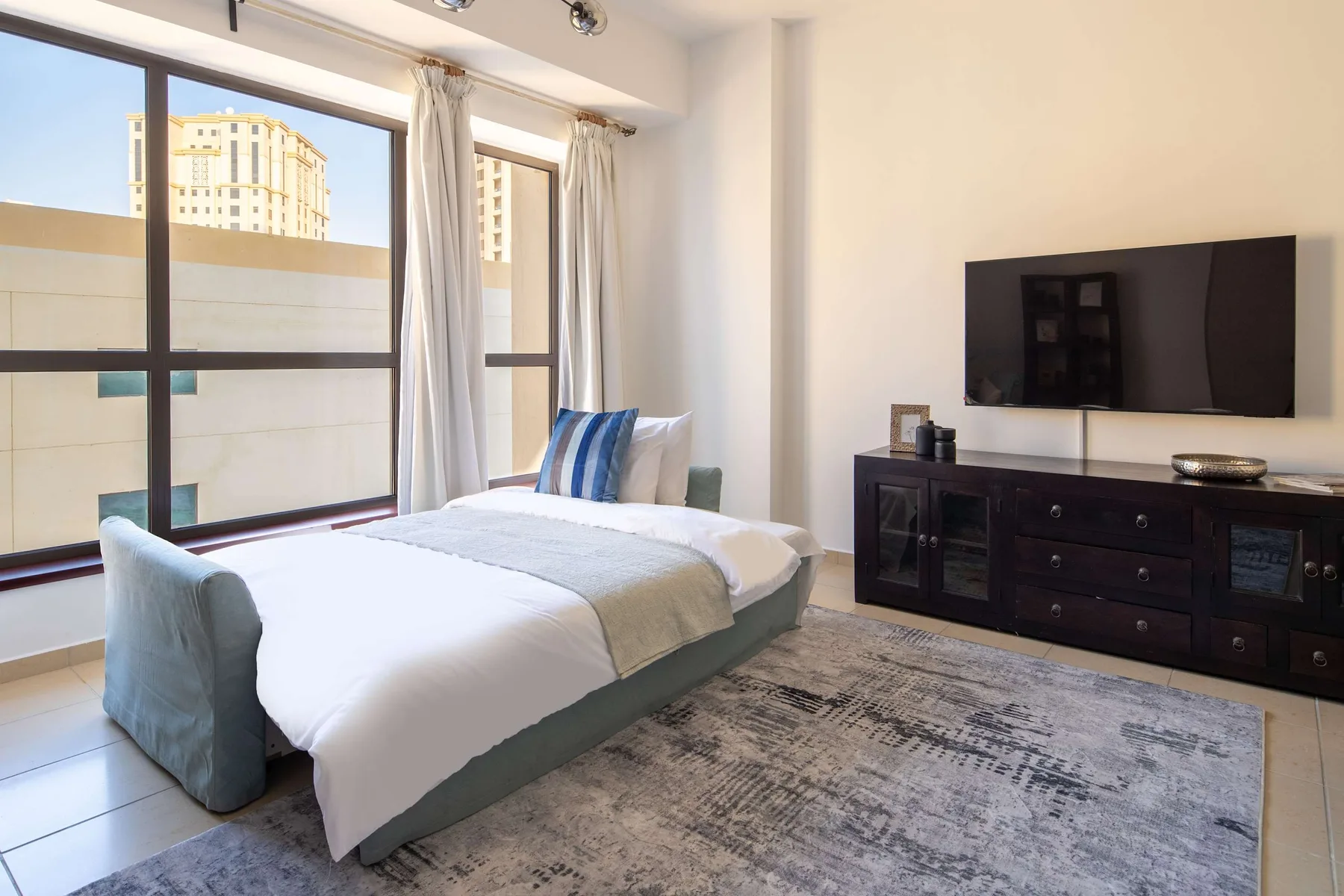 Un dormitorio luminoso con una cama tendida junto a grandes ventanas y un televisor.