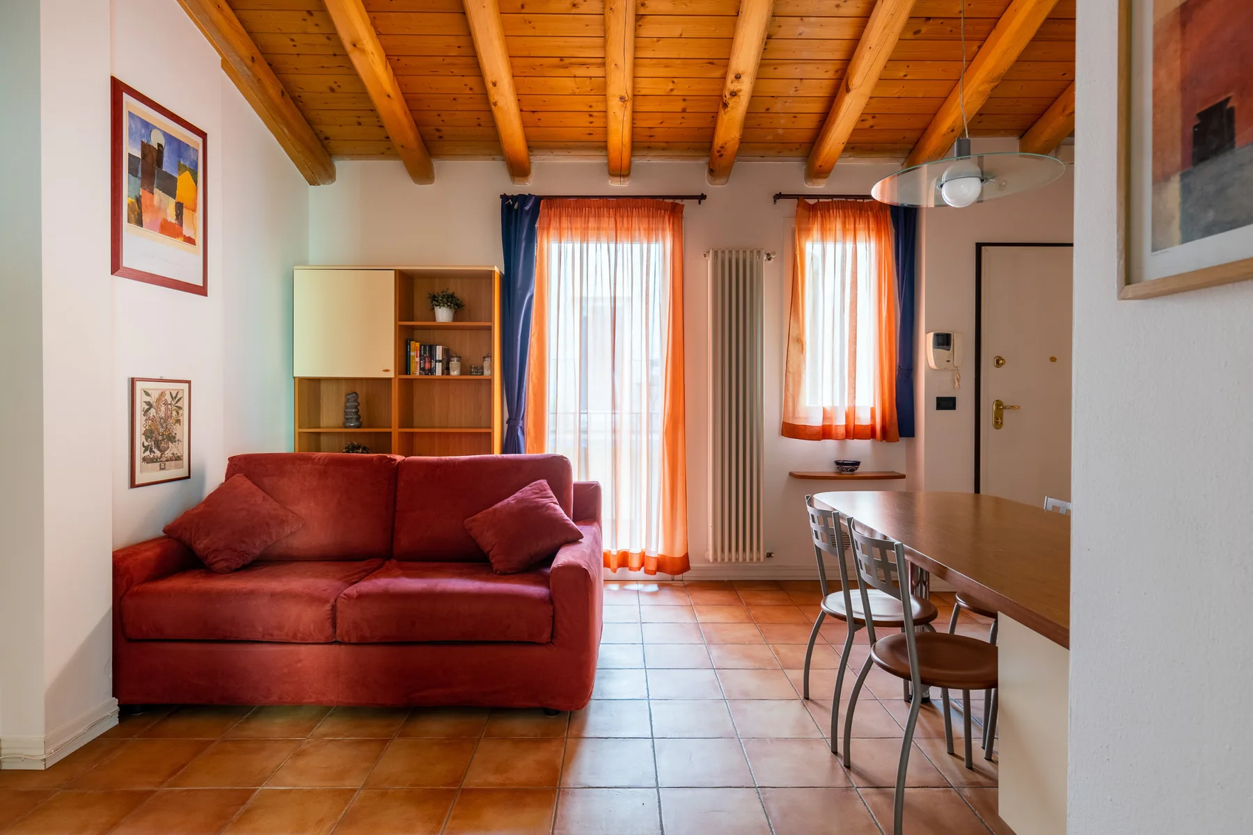 Una zona de estar y comedor con asientos rojos, una mesa y ventanas altas con cortinas.
