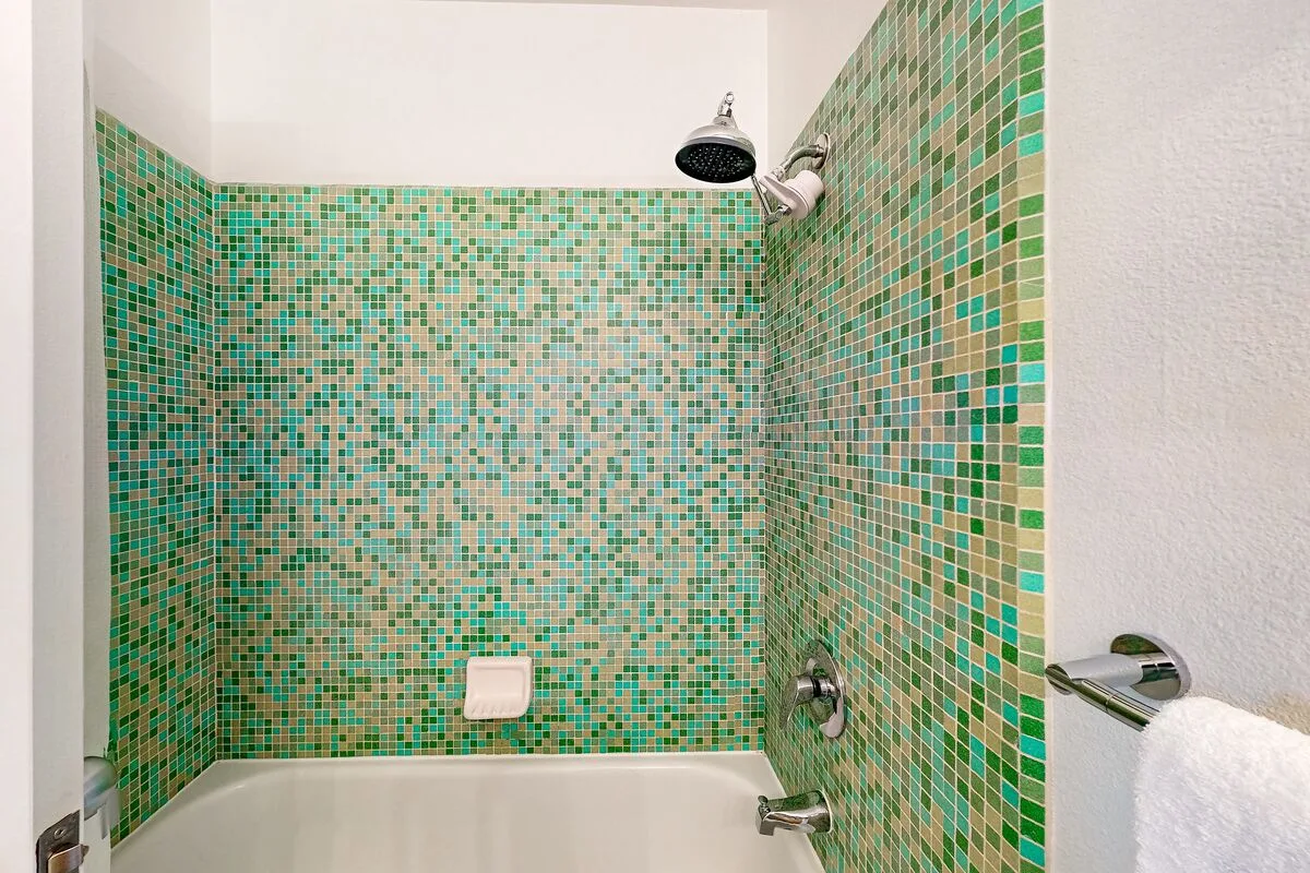 Una bañera está bajo una ducha, junto a paredes cubiertas de mosaicos verdes.