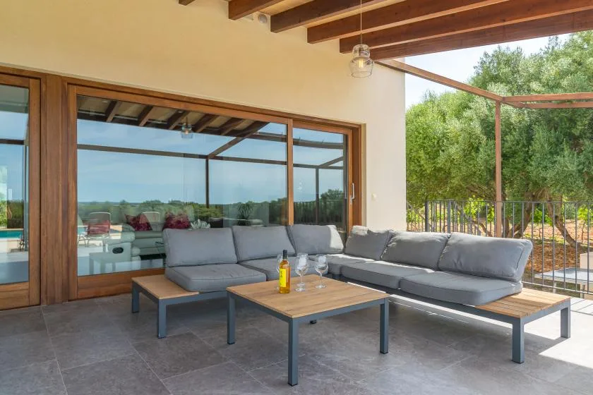 A covered outdoor lounge with gray seating and glass doors overlooking greenery.