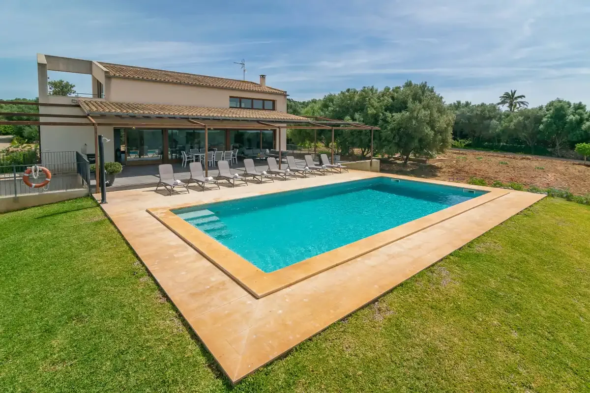 A modern house overlooks a swimming pool surrounded by a sunlit patio and lawn.
