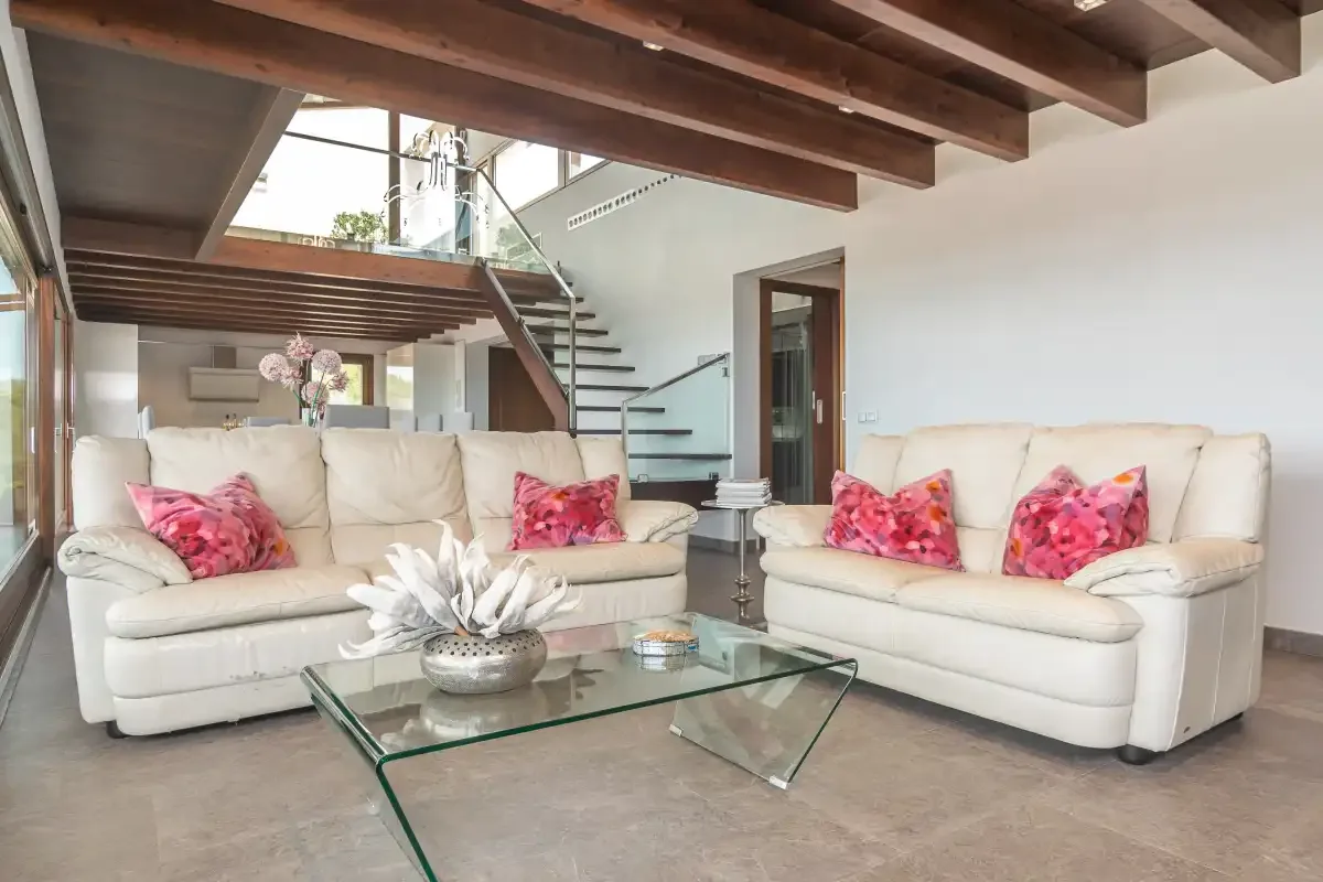 A bright living room with cream seating and a glass coffee table beneath exposed beams.