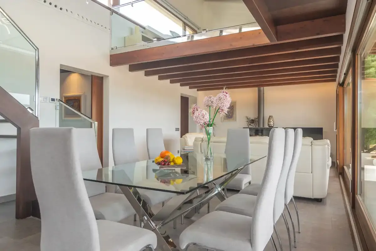 A dining area with a glass table and white seating beneath exposed ceiling beams.