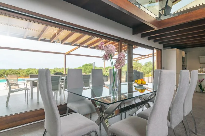 A dining area with a glass table and tall upholstered seating beside open patio doors.