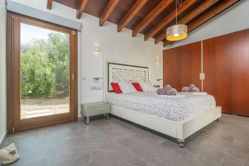 A bedroom with a white bed, wooden ceiling beams, and a glass door opening to greenery.