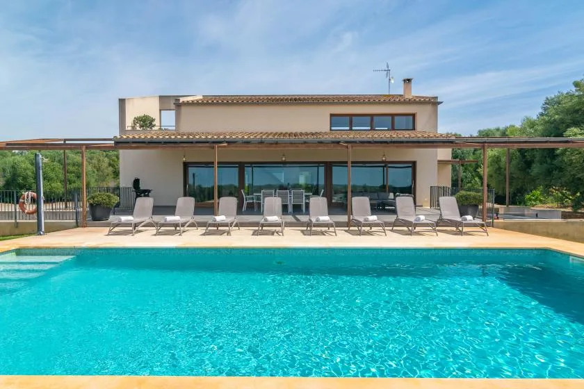 A modern house overlooks a swimming pool, with outdoor seating beside the water.
