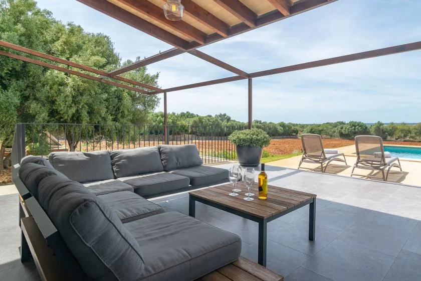A covered outdoor seating area with a sectional sofa, coffee table, and poolside loungers.