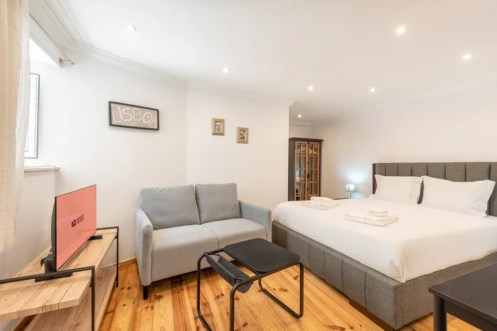 A bedroom with a large bed, gray seating, and light wood flooring.