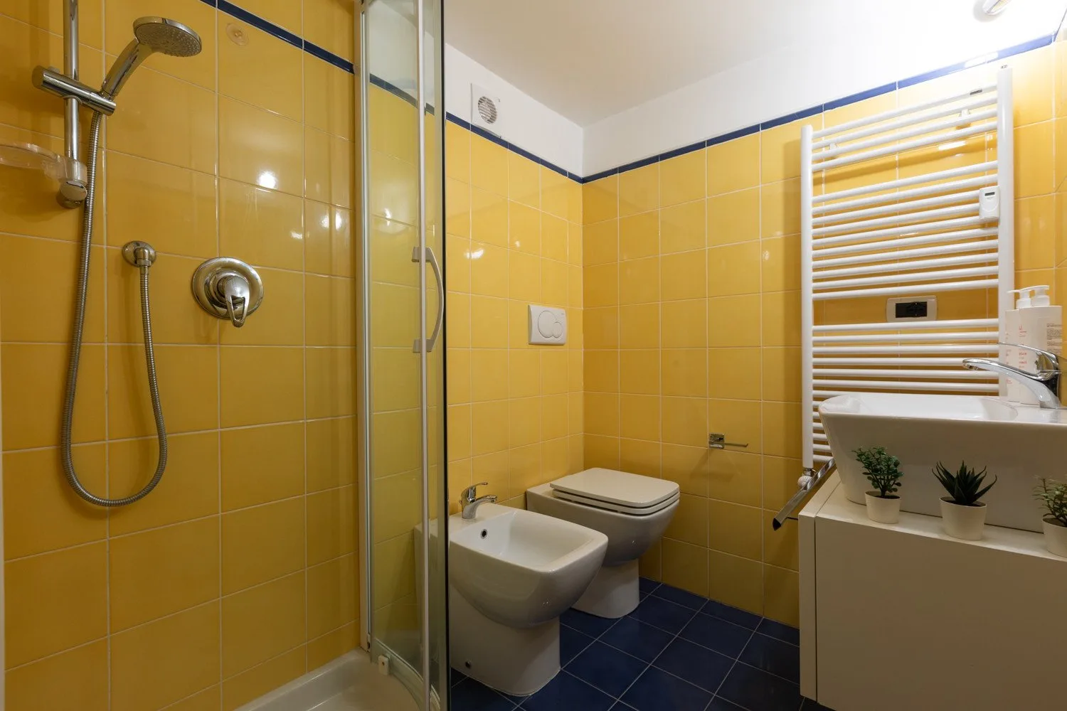 Un baño con azulejos amarillos, ducha de vidrio y accesorios blancos.