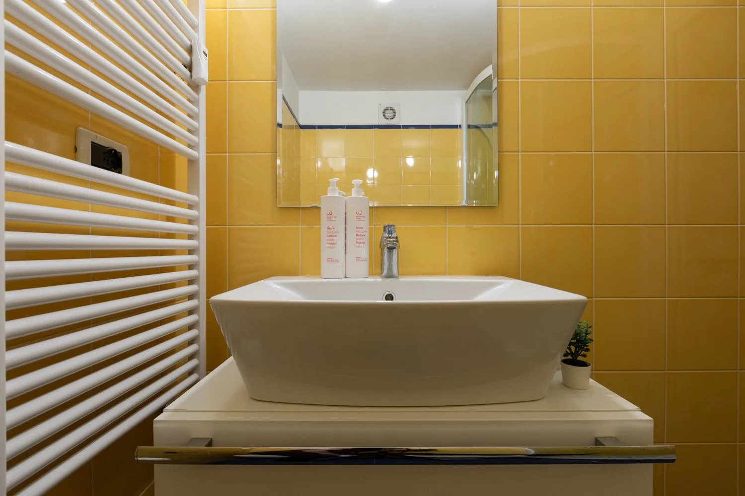 Un lavabo de baño está bajo un espejo, frente a paredes de azulejos amarillos.