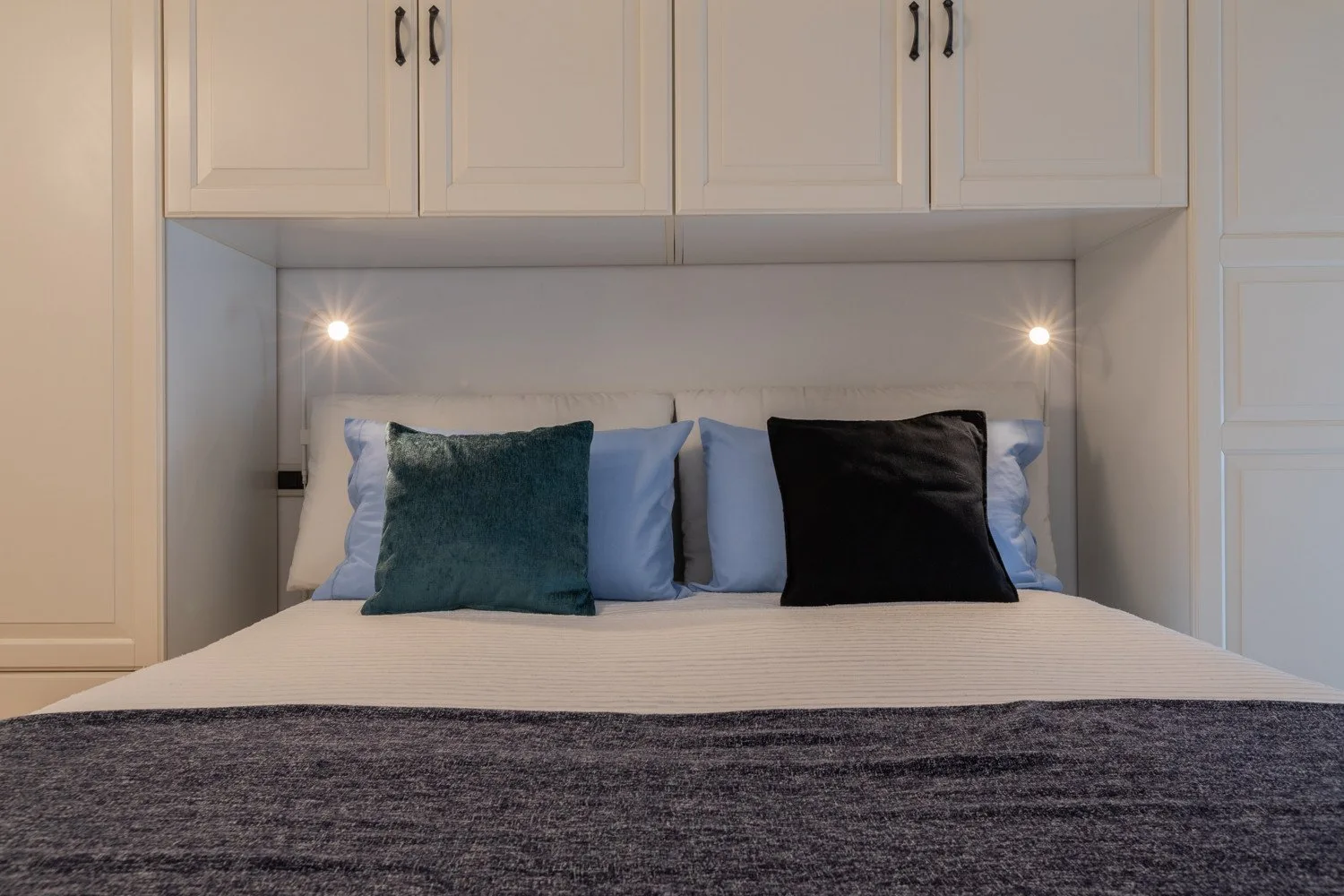 Una cama bien arreglada está bajo armarios empotrados, con cojines decorativos y luces junto a la cama.