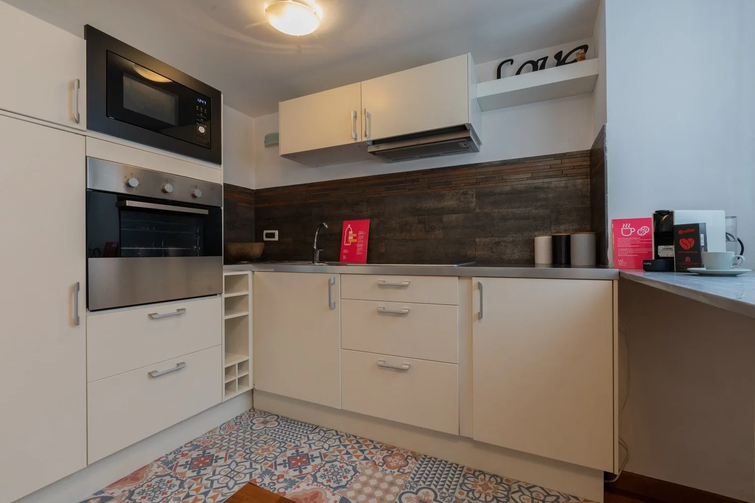 Una cocina compacta con gabinetes blancos, electrodomésticos integrados y un piso de baldosas estampadas.