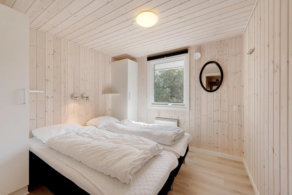 A bright bedroom with two beds, a window, and pale wood-paneled walls.