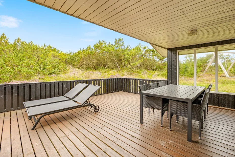 A covered outdoor deck has lounge seating and a dining table overlooking greenery.