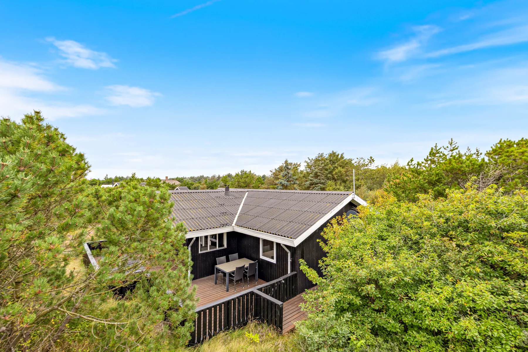 A dark wooden vacation house sits among leafy trees under a blue sky.