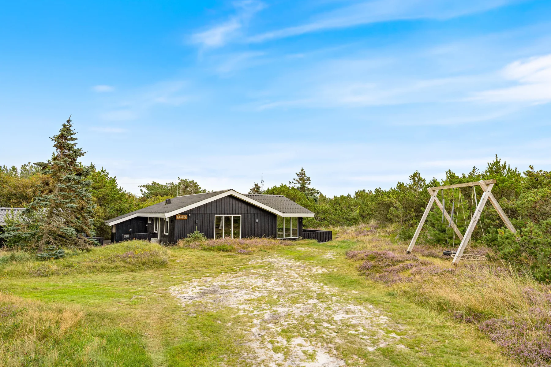 A dark house stands beside a sandy path in a grassy, shrub-lined landscape.